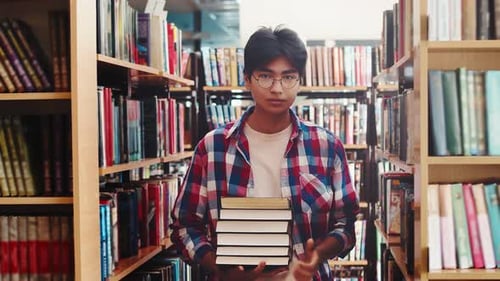 Young Male Student of Asian Appearance is Standing in the Library with Books Among the Shelves Turns