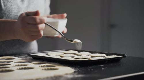 Woman carefully shakes sugar from a spoon onto mince pies in a baking tin to prepare for oven. Lady
