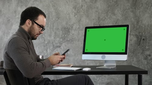 Man Uses Phone at Desk with Green Screen Computer