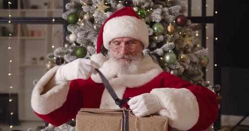 Santa Claus unwraps gift in front of Christmas tree