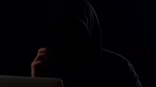 Hooded Figure Working on Laptop in Dark Room