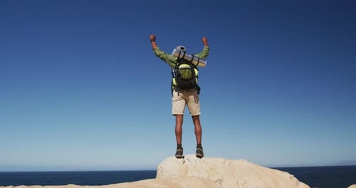 African american man hiking in mountains standing on rock raising his hands by the sea