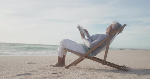 Latin senior hispanic woman relaxing on sunbed on beach at sunset, reading book