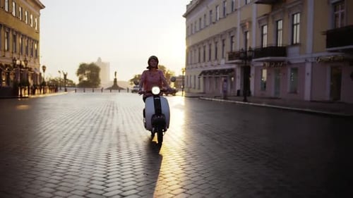 Junges schönes Mädchen fährt bei Sonnenaufgang mit dem Roller durch die Stadt
