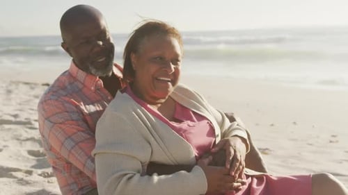 Smiling senior african american couple embracing and sitting on sunny beach