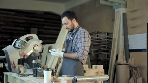 Carpenter with Beard Makes Something on His Smart Phone in Workshop.
