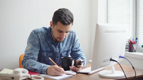 Junger Geschäftsmann sitzt in einem stilvollen, modernen Büro am Computer und macht sich mit seinem Stift Notizen