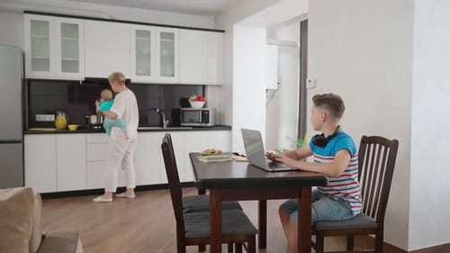 Mother and Infant in Kitchen, Teenager Using Laptop