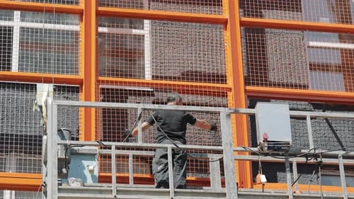 Ein Baumeister in einem Aufzug installiert ein schützendes Metallgitter an der Fassade des Hauses
