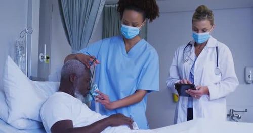 Diverse female doctors putting oxygen mask on male patient in hospital bed all wearing face masks