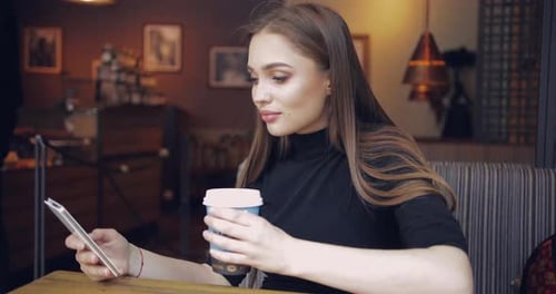 Young Beautiful Girl Sitting at Table in Cafe with Cup of Coffee in Hands and Browsing Smartphone