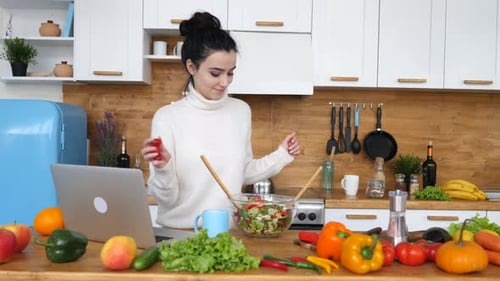 Young Woman Mixing Salad with Laptop in Kitchen