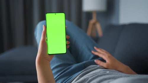 Woman at Home Lying on a Sofa and Using Smartphone with Green Mock-up Screen in Vertical Mode. Girl