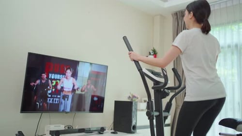 Hermosa joven asiática que usa bicicleta estática, sigue las instrucciones del video del entrenador en línea.