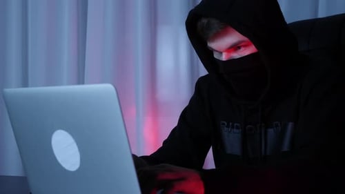 Hacker face in black mask and hood looking at laptop screen sitting in dark room. Online crimes