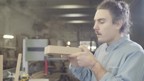 Caucasian Woodworker Blowing Sawdust off Piece of Wood in Basement Workshop