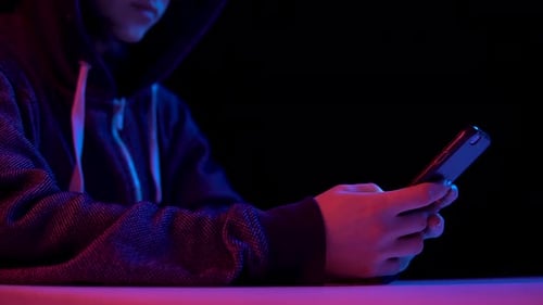 Young Woman in a Hood with a Smartphone. Hacker Makes a Hack Through the Phone. Blue and Red Light