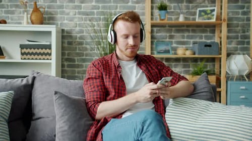 Slow Motion of Joyful Guy in Headphones Listening To Music and Using Smart Phone