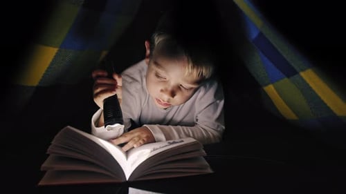 Boy reading a book with a flashlight under the covers at night