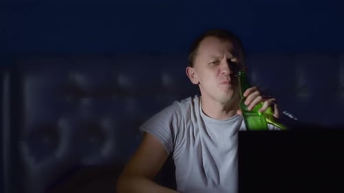 Cheerful man with a bottle of beer sits at the TV at night, raises his hand up, camera movement