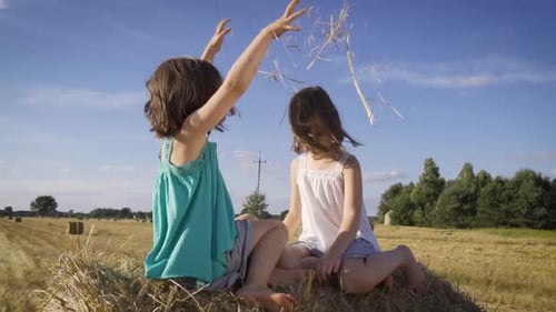 Two Charming Girls Are Sitting on a Roll of Mown Rye in a Field