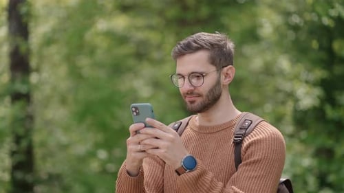 A Young Man with a Backpack in the Woods in Nature Writes Messages on a Mobile Phone