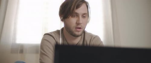 Ein junger Mann spricht über Kopfhörer mit jemandem, während er an seinem Laptop arbeitet.