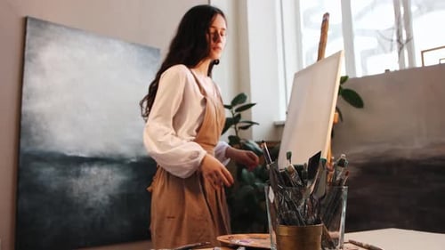 Art Studio Young Woman Putting a Canvas on an Easel and Stands in the Artist Pose