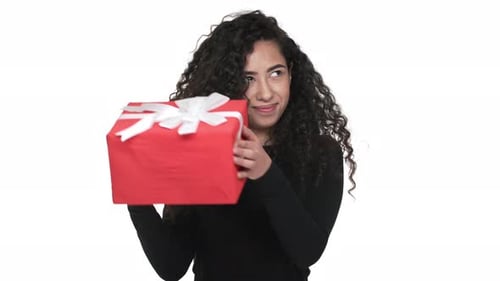 Retrato de una curiosa mujer de 20 años sacudiendo una caja roja envuelta para regalo emocionada y sorprendida