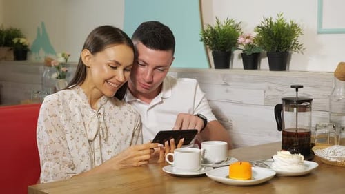 Girl and Guy Looks at Mobile Phone at Table in Sweet Shop