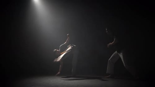 Beaux hommes pratiquant la capoeira dans l'obscurité contre les projecteurs en studio