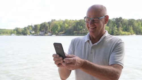 Happy Senior Man Using Phone By The Lake