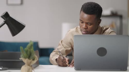 Man Writes at Desk Near Laptop