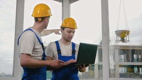 Two builders wearing protective helmets working on a laptop