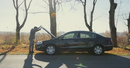 Besorgter Mann, der seinen Kopf mit der Hand hält, steht neben seinem kaputten Auto mit angehobener Motorhaube auf der Straße