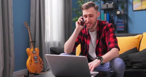 Young Man Using Laptop and Talking on Phone at Home