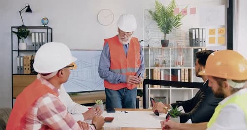 Ingeniero con casco y chaleco explicando el proyecto de construcción representado en una pantalla digital y una pared de vidrio