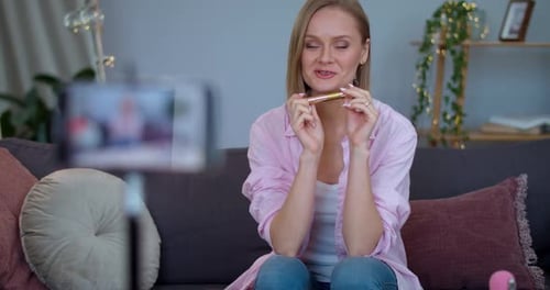 Atractiva vloguera haciendo una reseña de vídeo sobre un producto de maquillaje mientras está sentada en el sofá. Joven