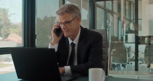 A businessman talking on the phone while working on a laptop at the office.