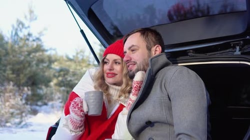 Young Couple a Man and a Woman are Sitting in the Trunk of a Car in a Winter Snowy Forest Hugging