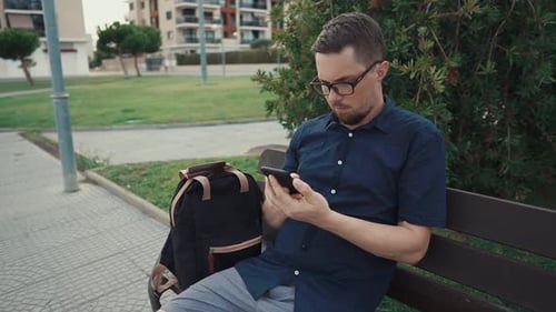 Serious Man Is Sitting on a Bench in Alley in City Park, Typing on Mobile Phone