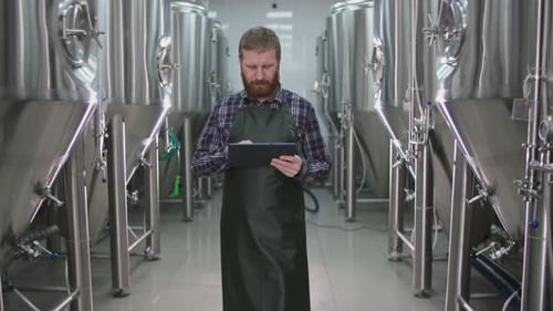 Ein männlicher Brauer mit Bart kommt an Biertanks vorbei und überprüft die Messwerte mit einer Tablette