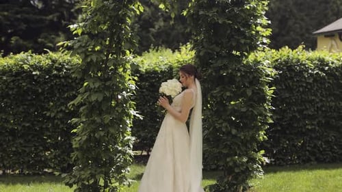 Beautiful Stylish Bride in White Wedding Dress and Veil Holding Wedding Bouquet in Hands in Park