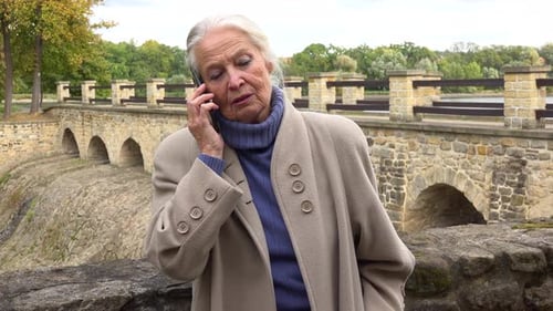 An Elderly Woman Leans Against the Stone Barrier of a Bridge and Talks on a Smartphone
