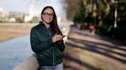 Portrait of a Beautiful Adult Woman with Glasses Standing on the Street in Spring with a Phone in