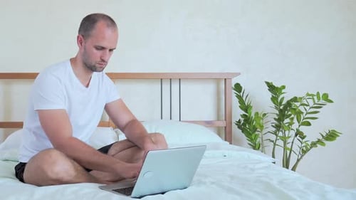 Man Works on Laptop Sitting on Bed Indoors