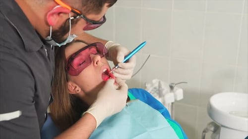 Doctor Orthodontist with Glasses Whitens the Teeth of a Young Woman with a Laser