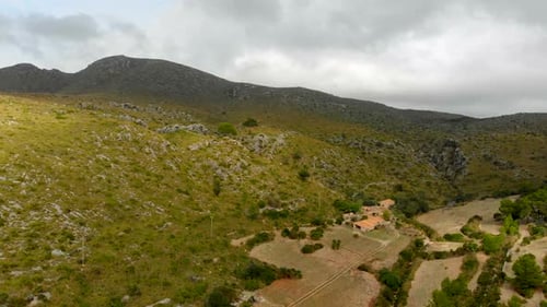 Drone footage moving towards the mountains with a green hill and some buildings in the foreground. F