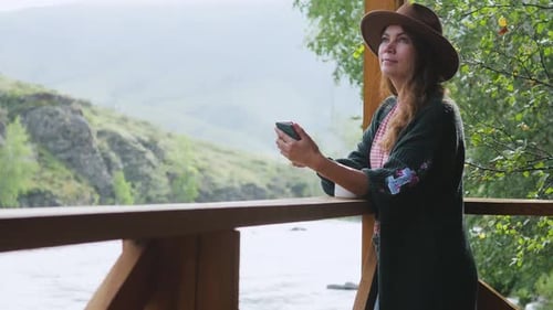 Woman Using Phone on Cabin Balcony Overlooking River