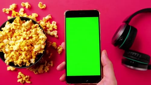 Man's hand holding smart phone with green touch screen and popcorn in a bowl on red background.